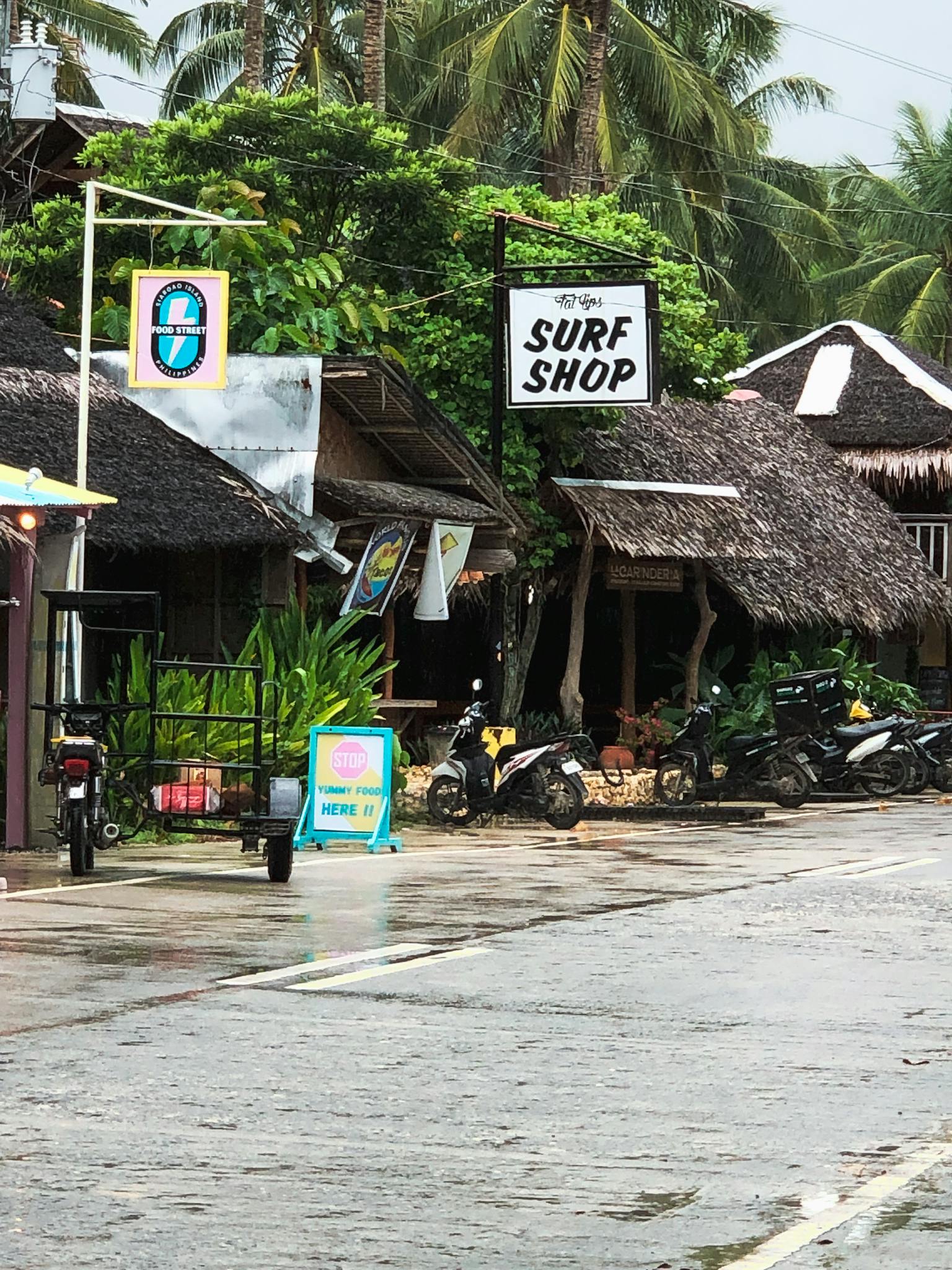 Cozy tropical surf shop surrounded by greenery and parked scooters on a rainy day.